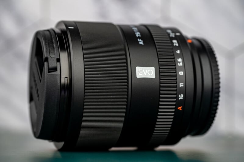 A close-up photo of a black camera lens with "EVO" branding, focus and aperture markings, and a lens cap, placed on a green surface against a blurred, geometric-patterned background.