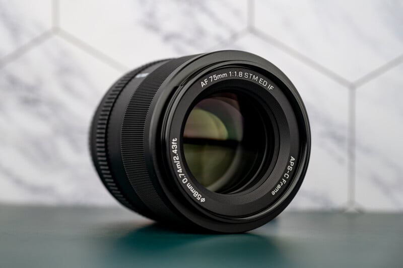 A black camera lens with white labelling sits on a teal surface in front of a white hexagonal tile background. The lens is marked "AF 75mm 1:1.8 STM ED IF" and has a large, reflective glass element.