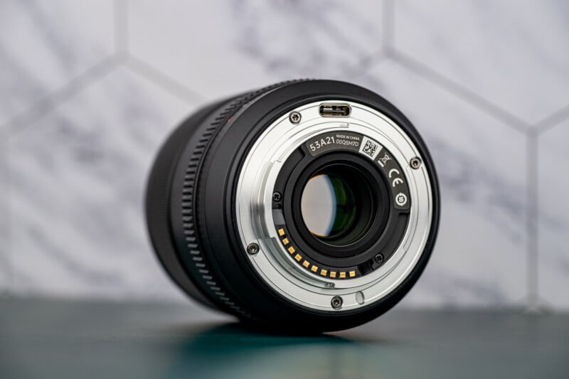 A close-up view of the rear side of a camera lens showing the metal mount, electronic contacts, and glass elements, placed on a green surface with a blurred geometric background.