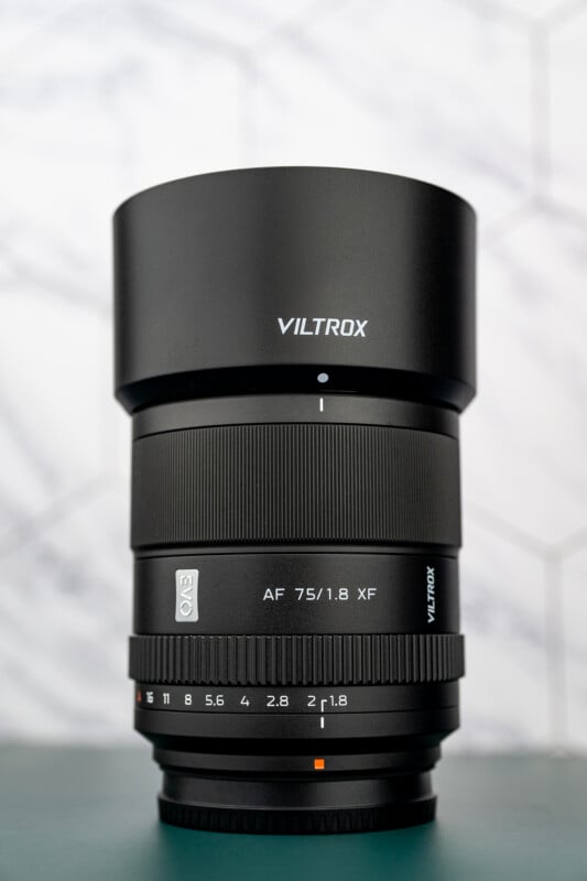 A close-up photo of a Viltrox AF 75mm f/1.8 XF camera lens standing upright on a green surface, with a geometric, light-colored background.
