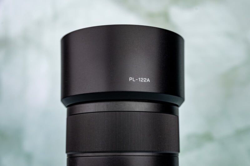A close-up of a black camera lens with "PL-122A" written on the lens hood, set against a blurred, light background.