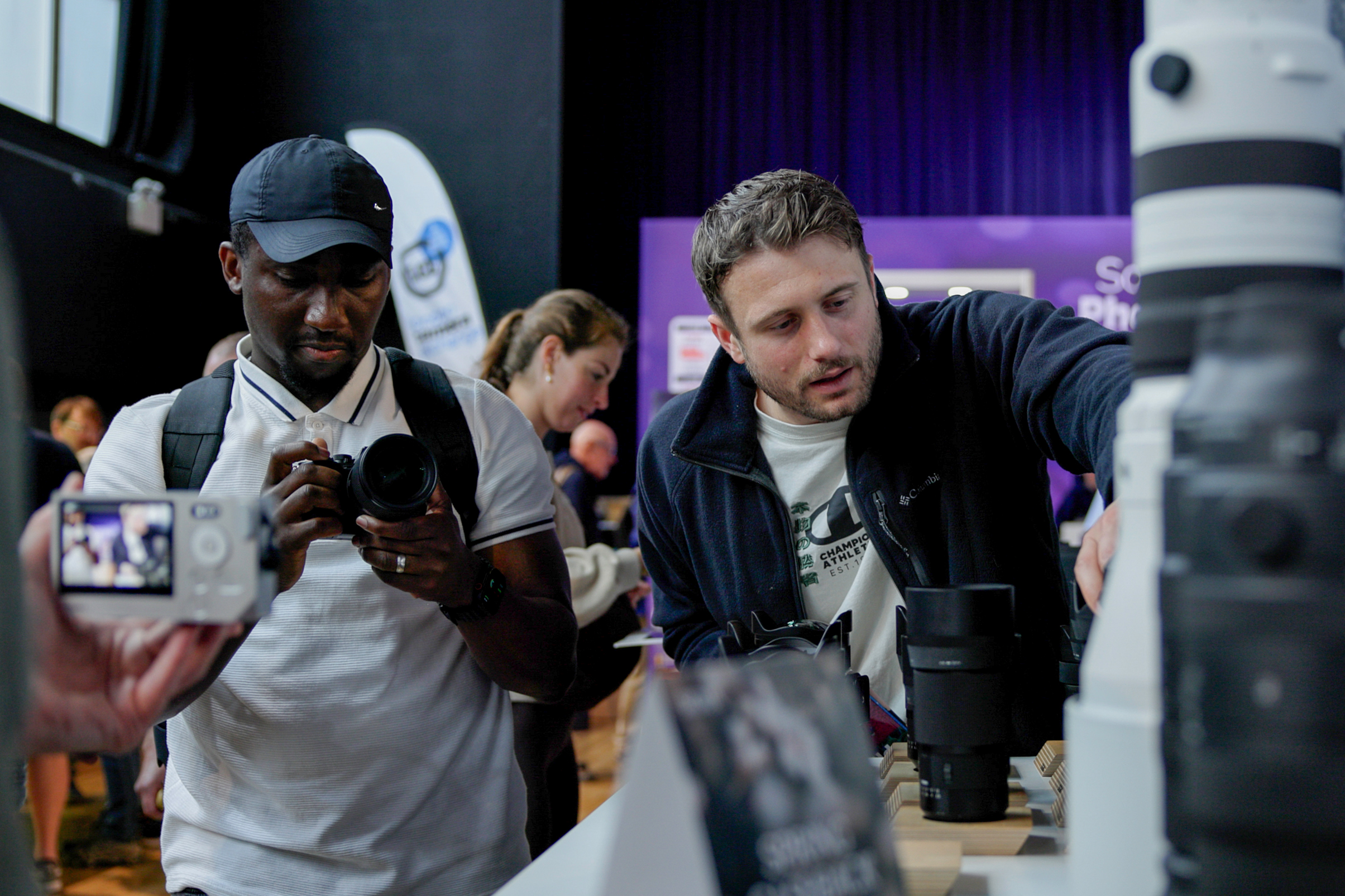 Two people examining cameras and lenses displayed on a table at an indoor event or exhibition