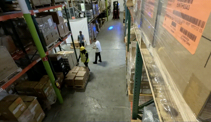 A video shows a corridor with shelves of boxes on either side and a pile of boxes on the ground. Three people are standing next to the pile of boxes. There’s a small explosion from one of the boxes on the floor, and it starts smoking.