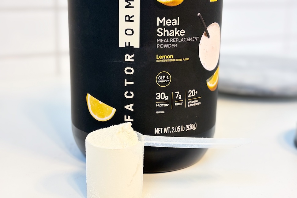 A canister of Facto Form Meal Replacement sitting on a countertop. A plastic scoop sits next to it, packed to the brim with off-white powder.