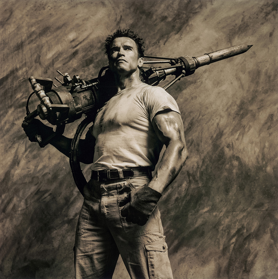 A low-angle black-and-white shot of Schwarzenegger in a polo shirt and trousers, arms raised wide with hands open, shot against a streaked sky that gives him an almost mythological scale.