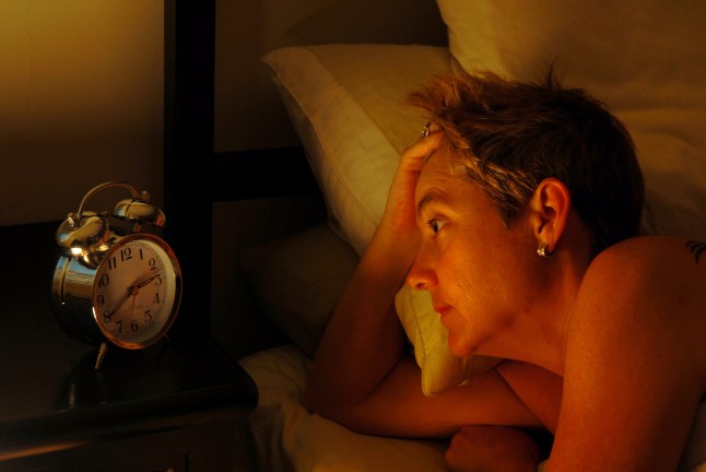Woman laying down in bed holding head looking at alarm clock