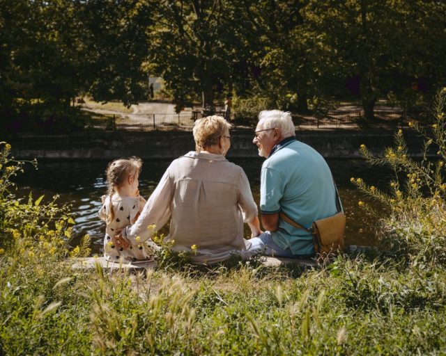 Rear view of grandparents sitting with granddaughter near pond at park - stock photo