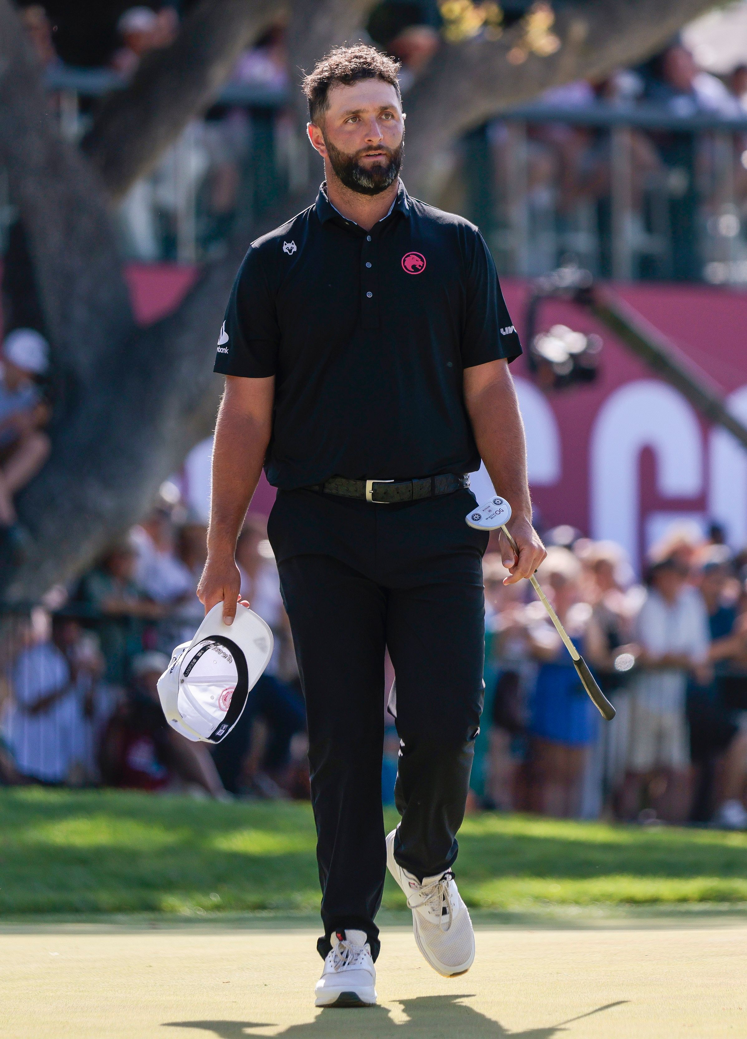 Male golfer in black outfit walks on green golf course, holding white hat.