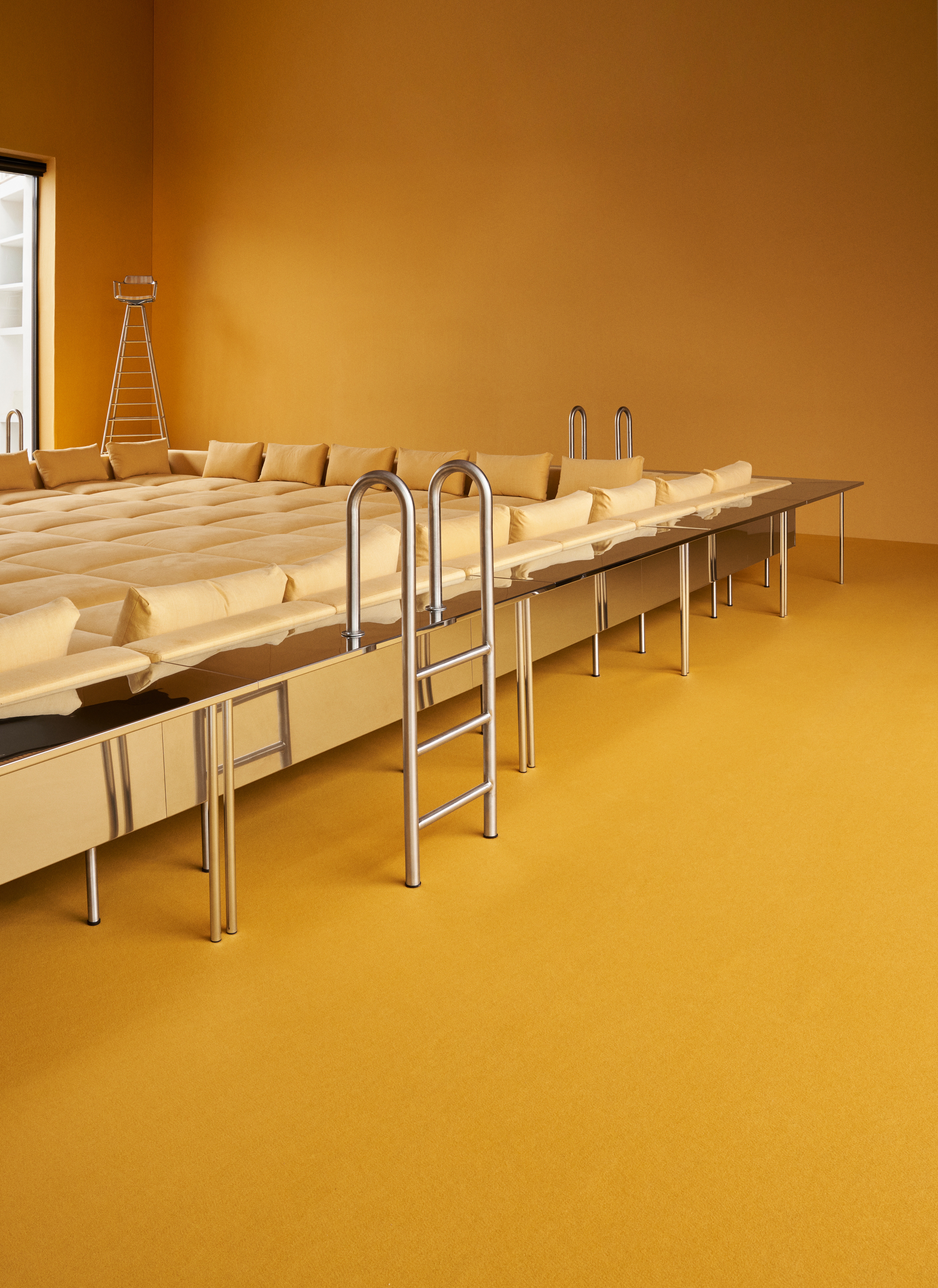 A playful installation comprising a chrome-clad, two-seat rocking chair, a butter yellow-painted kiosk, and a massive modular sofa with a square surface, butter yellow fabric, and chrome detailing.