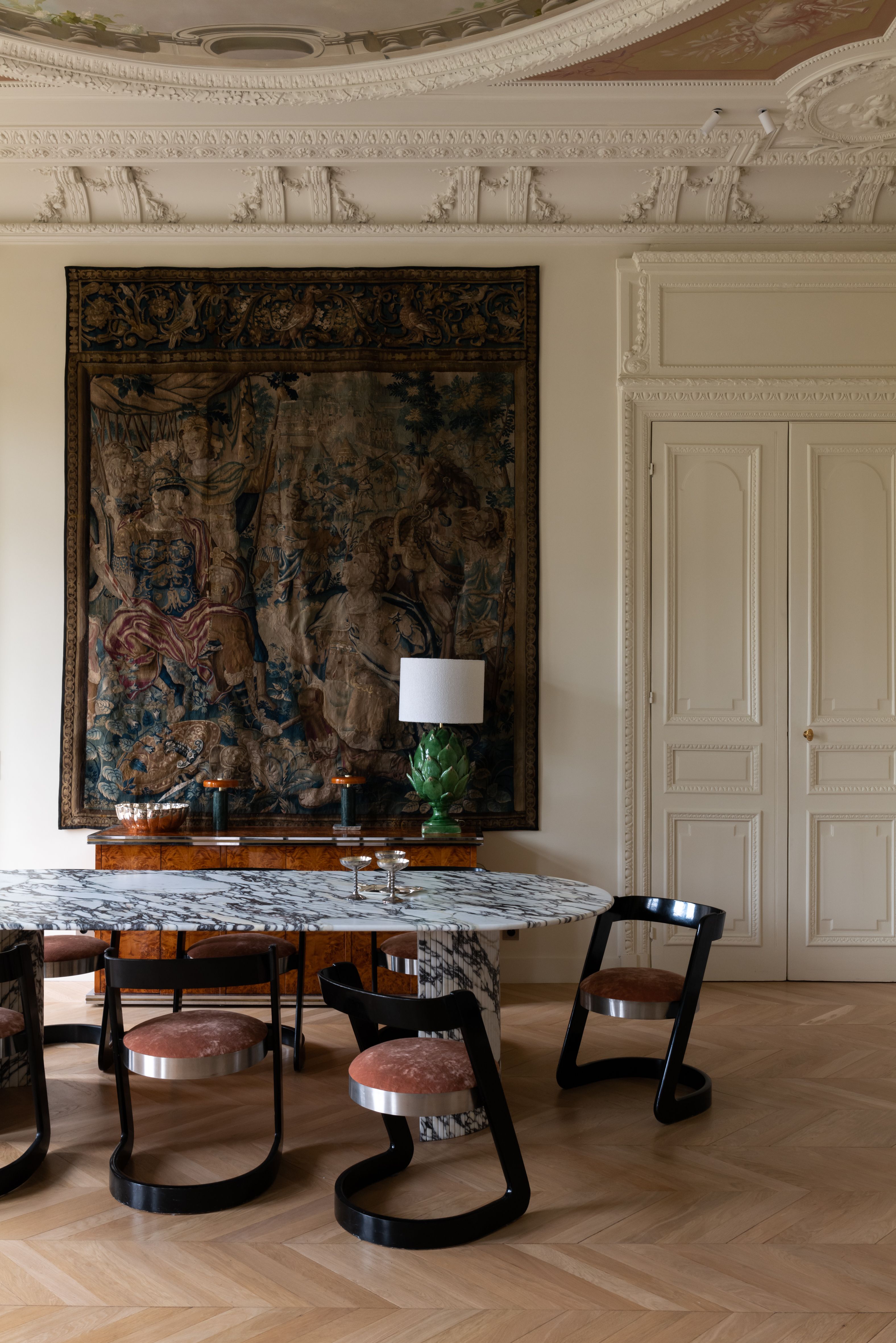 A grand dining room with a marble oval table and statement dining chairs with black backs and legs, and round seating with a large, dark tapestry hanging on an off-white white wall