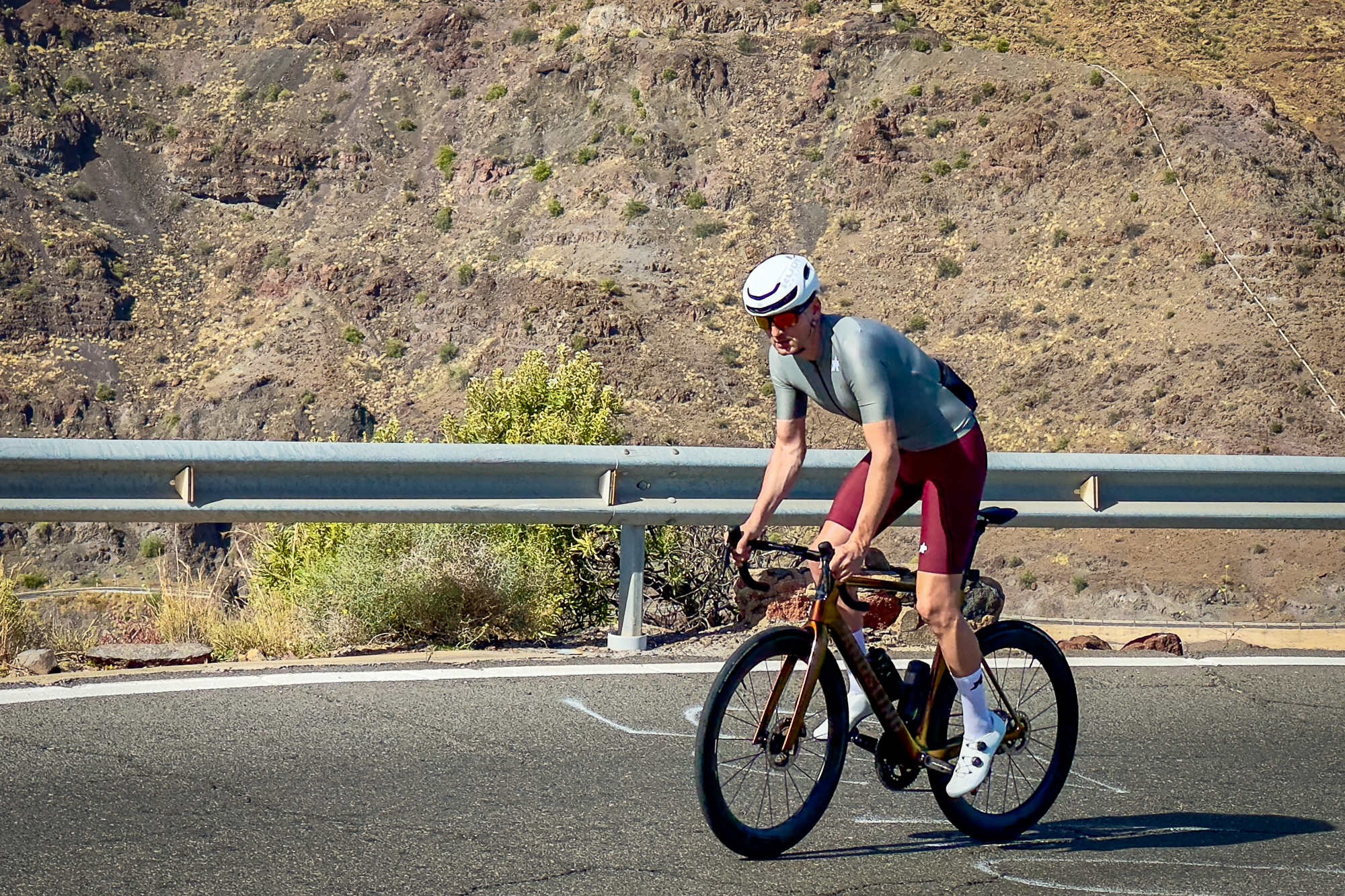 Man wearing green jersey, red shorts and white helmet riding out of the saddle up a hill