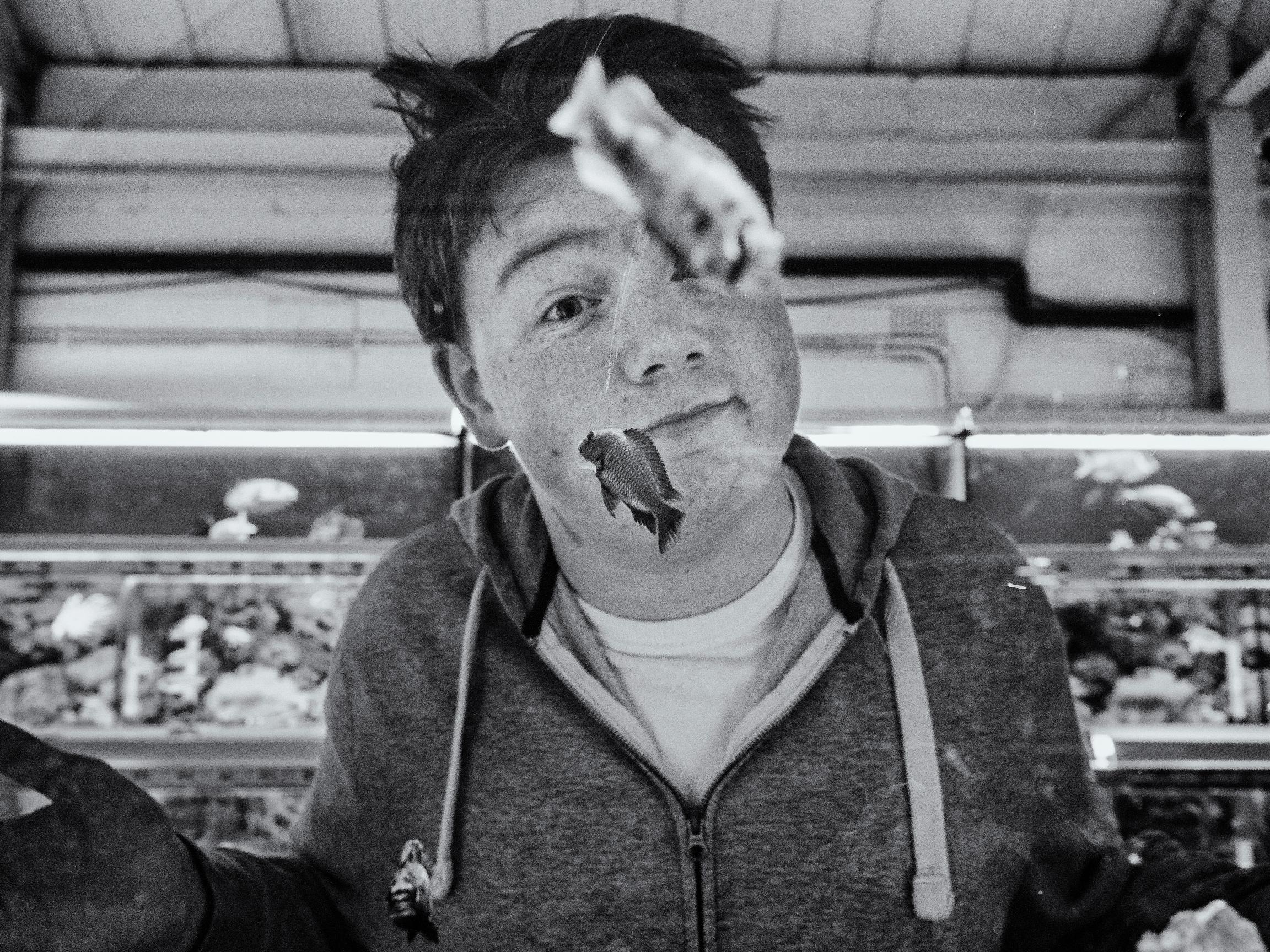 A black and white photo of a young man looking through the glass of a fish tank, with small fish appearing to swim directly in front of his face.