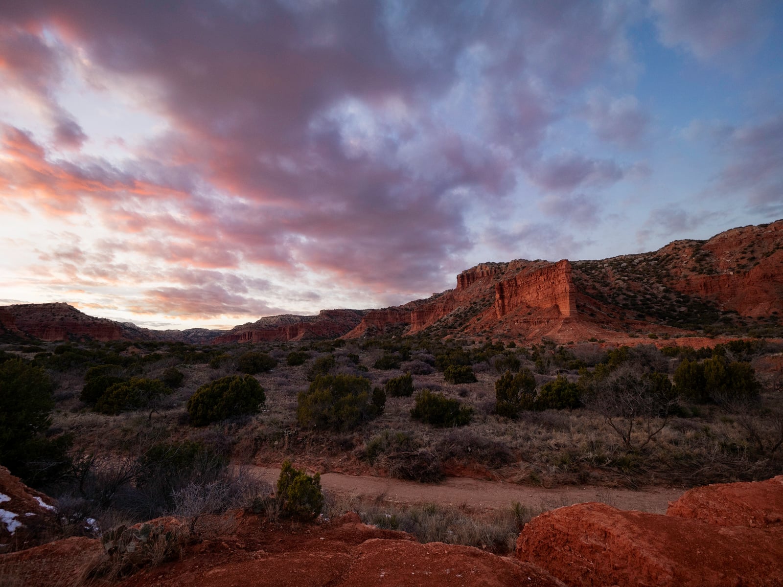 Red rock formations and green shrubs under a dramatic, colorful sunset sky with clouds, in a rugged, semi-arid landscape. A dirt path winds through the foreground.