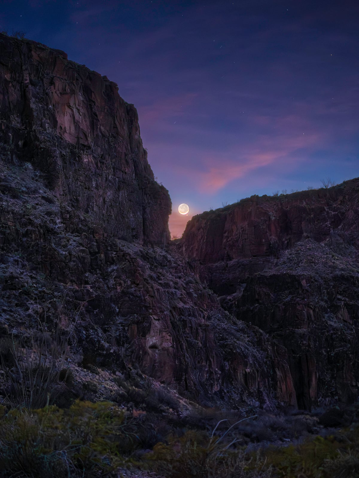 A crescent moon rises between two rocky canyon cliffs under a twilight sky, with faint stars and soft purple and pink clouds. Desert vegetation is visible in the foreground.