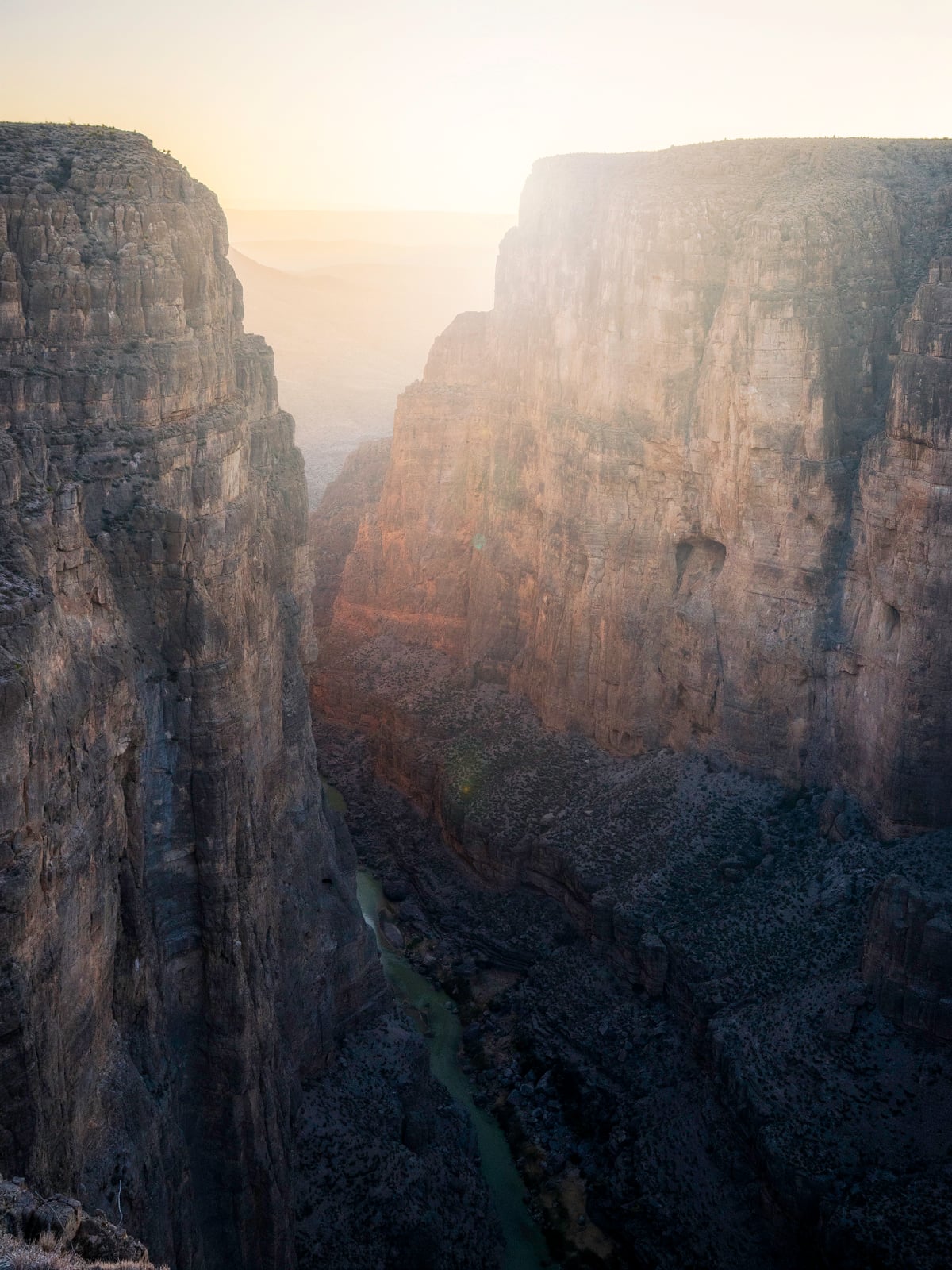 Sunlight filters through tall, rugged canyon walls with a narrow river winding along the canyon floor, casting dramatic shadows over the rocky landscape below.