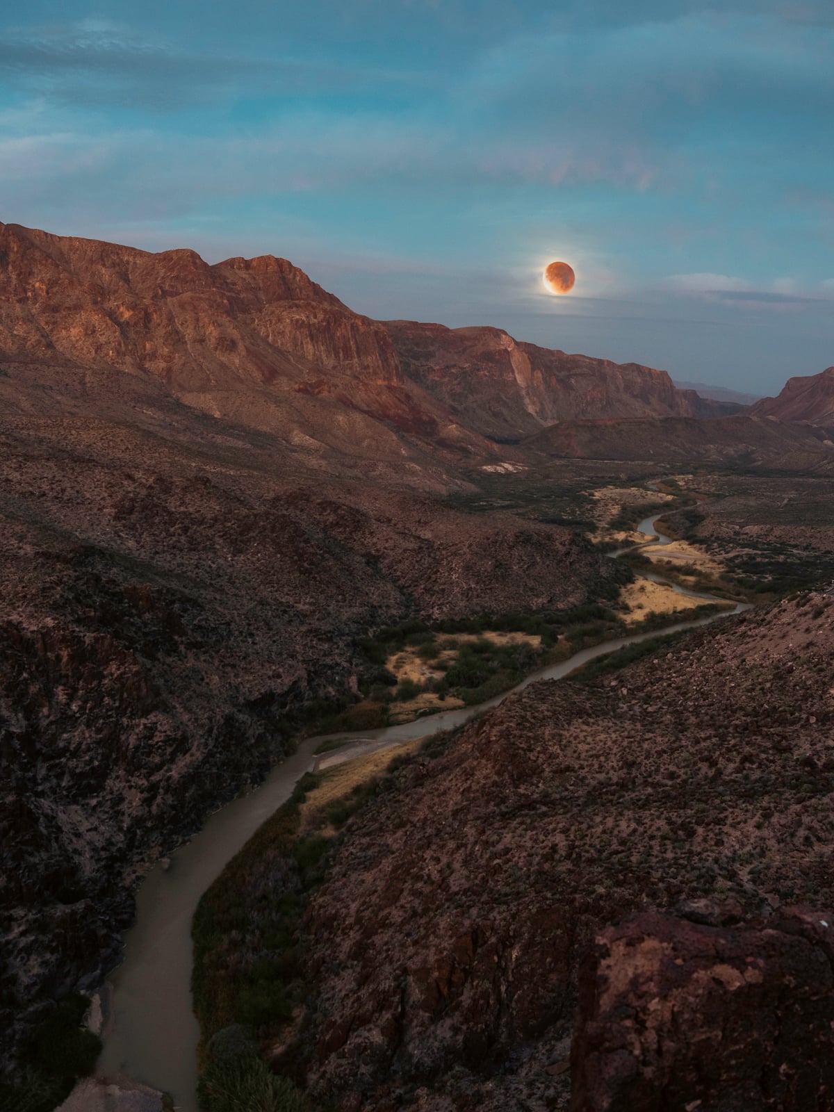 A winding river flows through a rocky desert canyon at dusk, with reddish mountains in the background and a partially eclipsed moon glowing above the horizon under a blue sky.