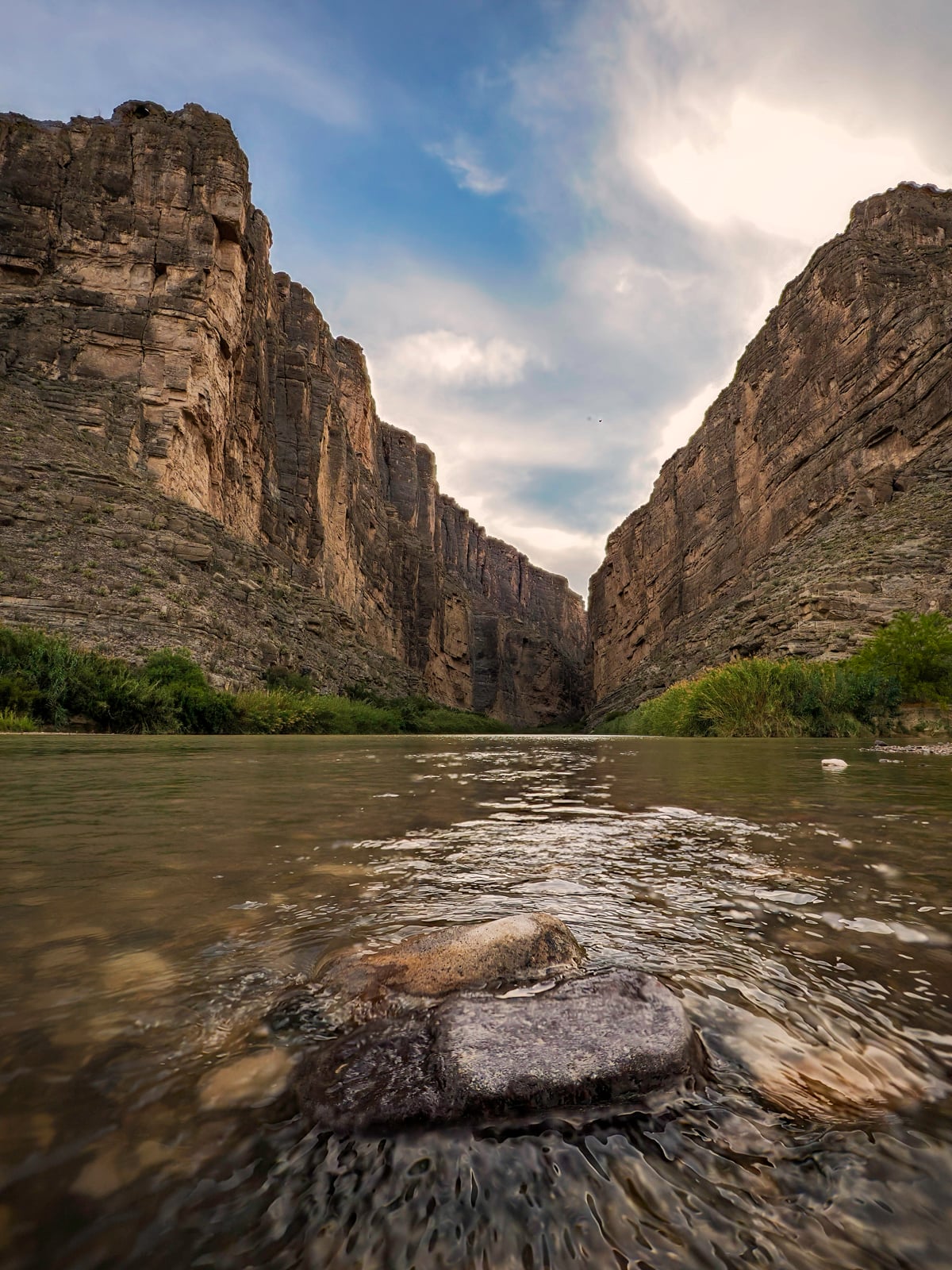 A river flows through a dramatic canyon with tall, steep rock walls on both sides, under a partly cloudy sky. Green shrubs grow along the riverbanks, and smooth stones are visible in the foreground.