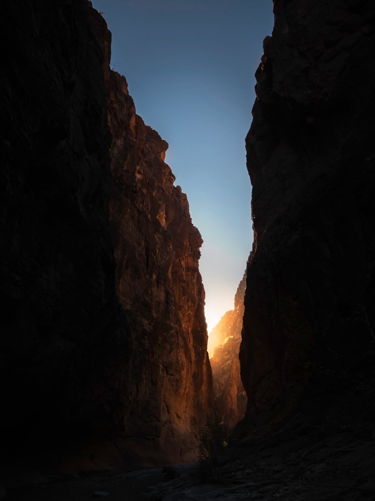 A narrow canyon with tall, dark rock walls. Warm sunlight glows between the cliffs, illuminating the rocks and sky at the canyon’s end. The foreground is mostly in shadow.