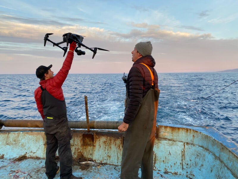 Two people in waterproof overalls and jackets stand on a weathered boat at sea; one holds a large drone overhead while the other looks on. The sky is cloudy with pastel colors, and the ocean extends to the horizon.