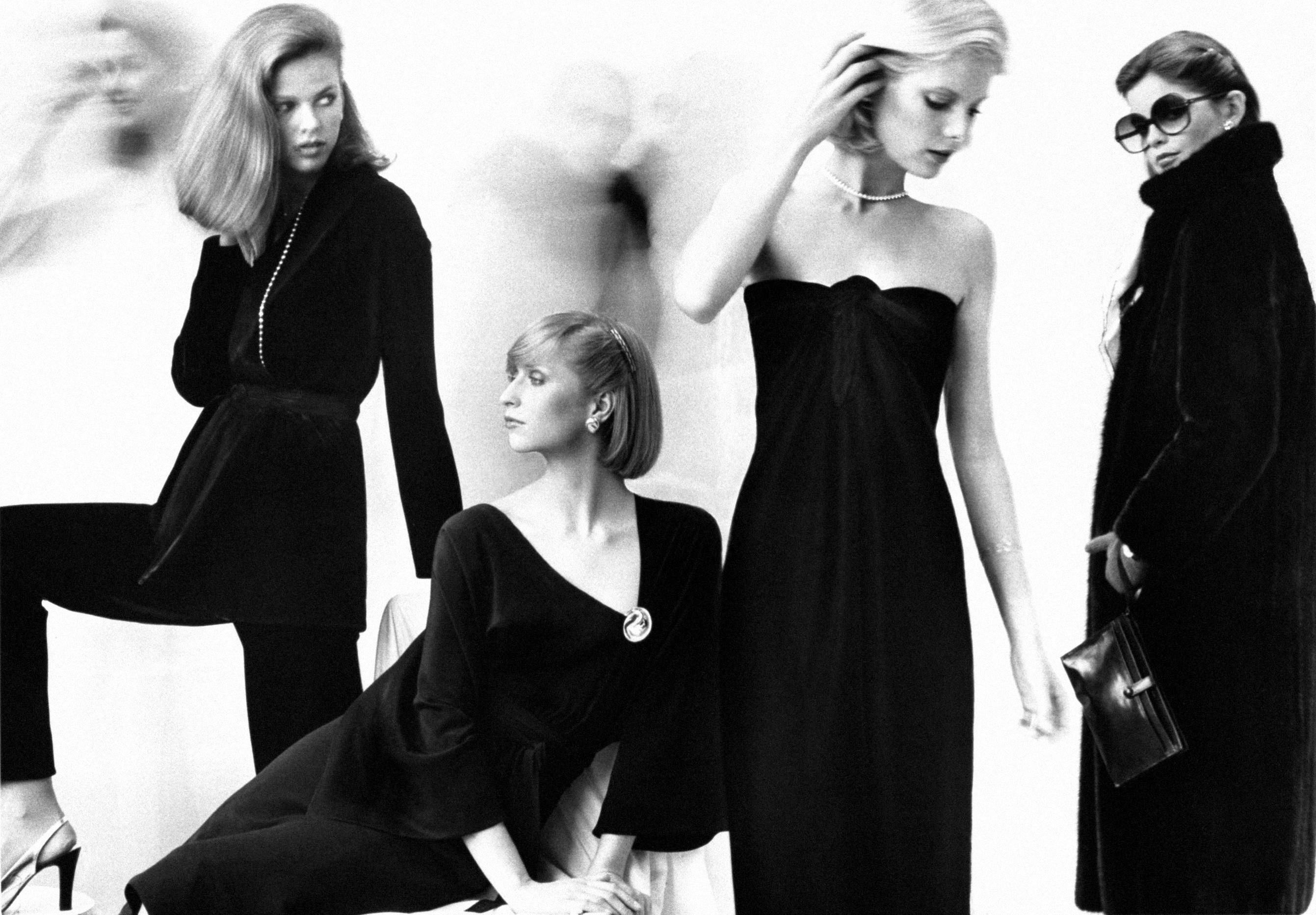 Four models in a dressing room, from left to right: Lisa Taylor wearing a velvet wrap jacket and matching pants; Chris Royer, seated, in a black silk crepe de chine dress with three-quarter sleeves, deep neckline and pin; Center model, standing, in a long black velvet strapless dress with diamond choker by Elsa Peretti; Right, wearing a dark mink coat by Halston, with handbag and large dark sunglasses. (Photo by Duane Michals/Conde Nast via Getty Images)