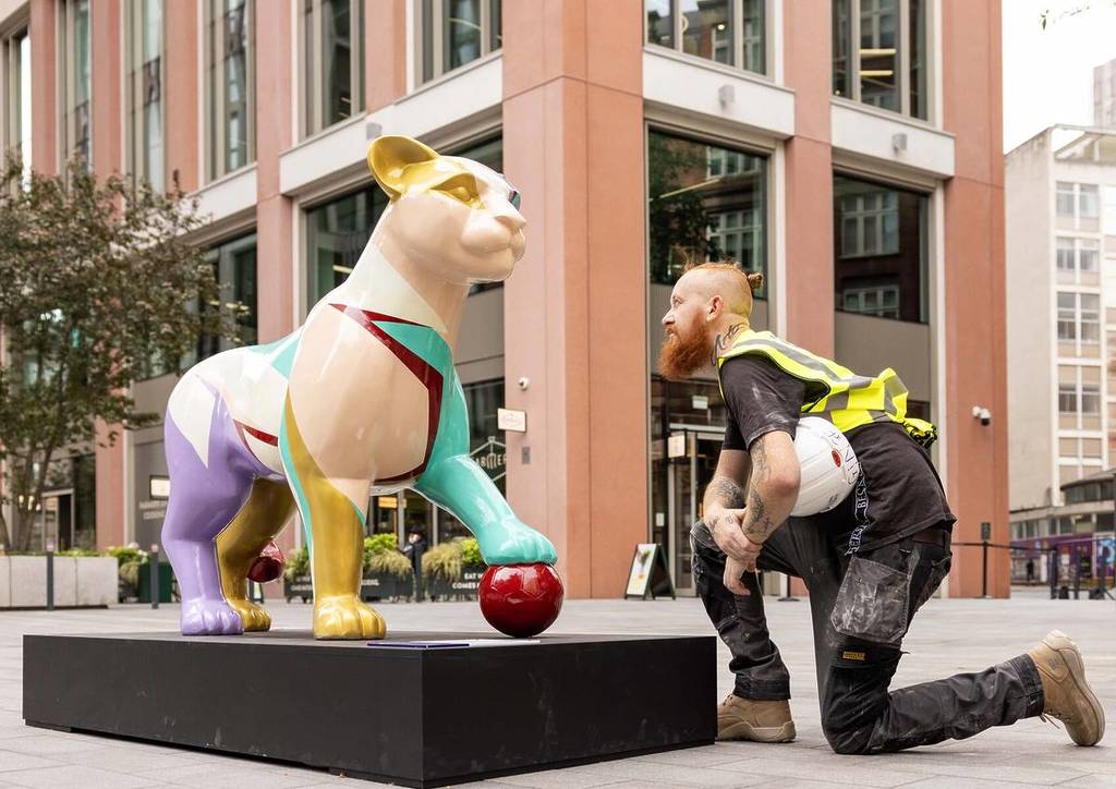 someone kneeling in front of and inspecting a sculpture of a lioness with a unique design, with one foot atop a football