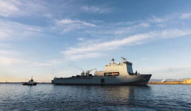 RFA Lyme Bay sails from Gibraltar escorted by a tugboat during a previous deployment, with calm water and blue skies surrounding the Bay-class support ship.
