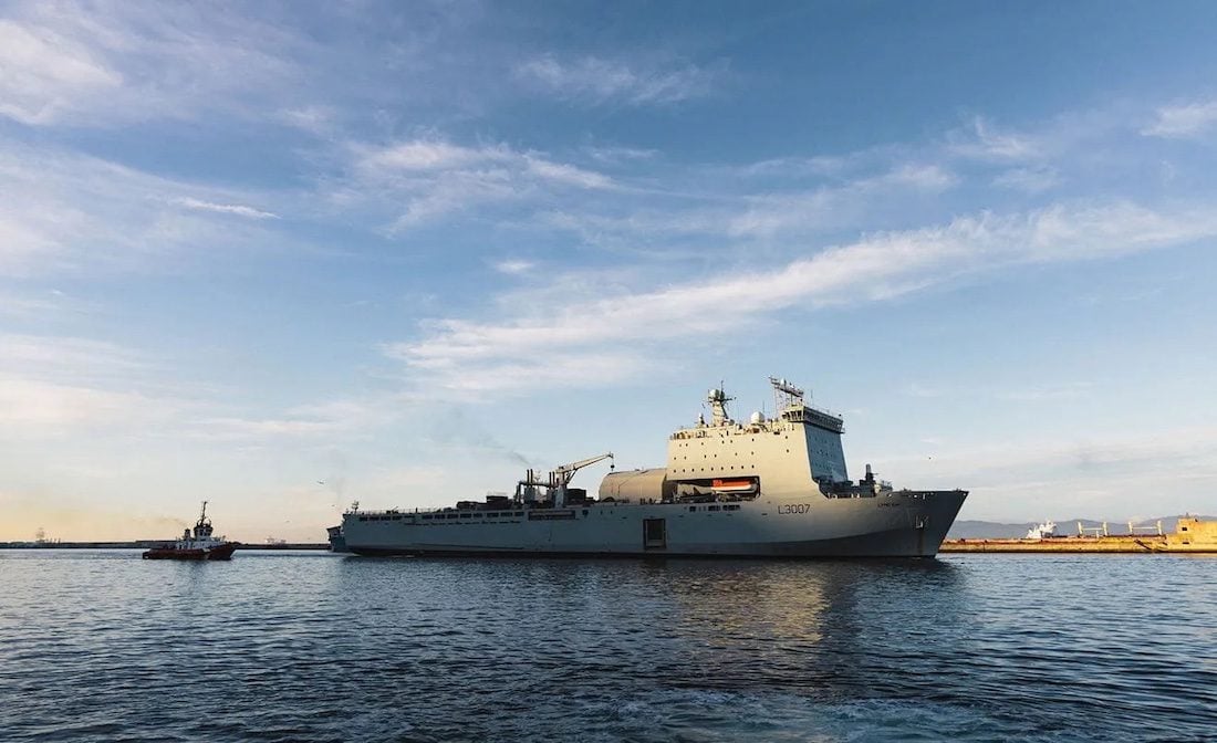 RFA Lyme Bay sails from Gibraltar escorted by a tugboat during a previous deployment, with calm water and blue skies surrounding the Bay-class support ship.