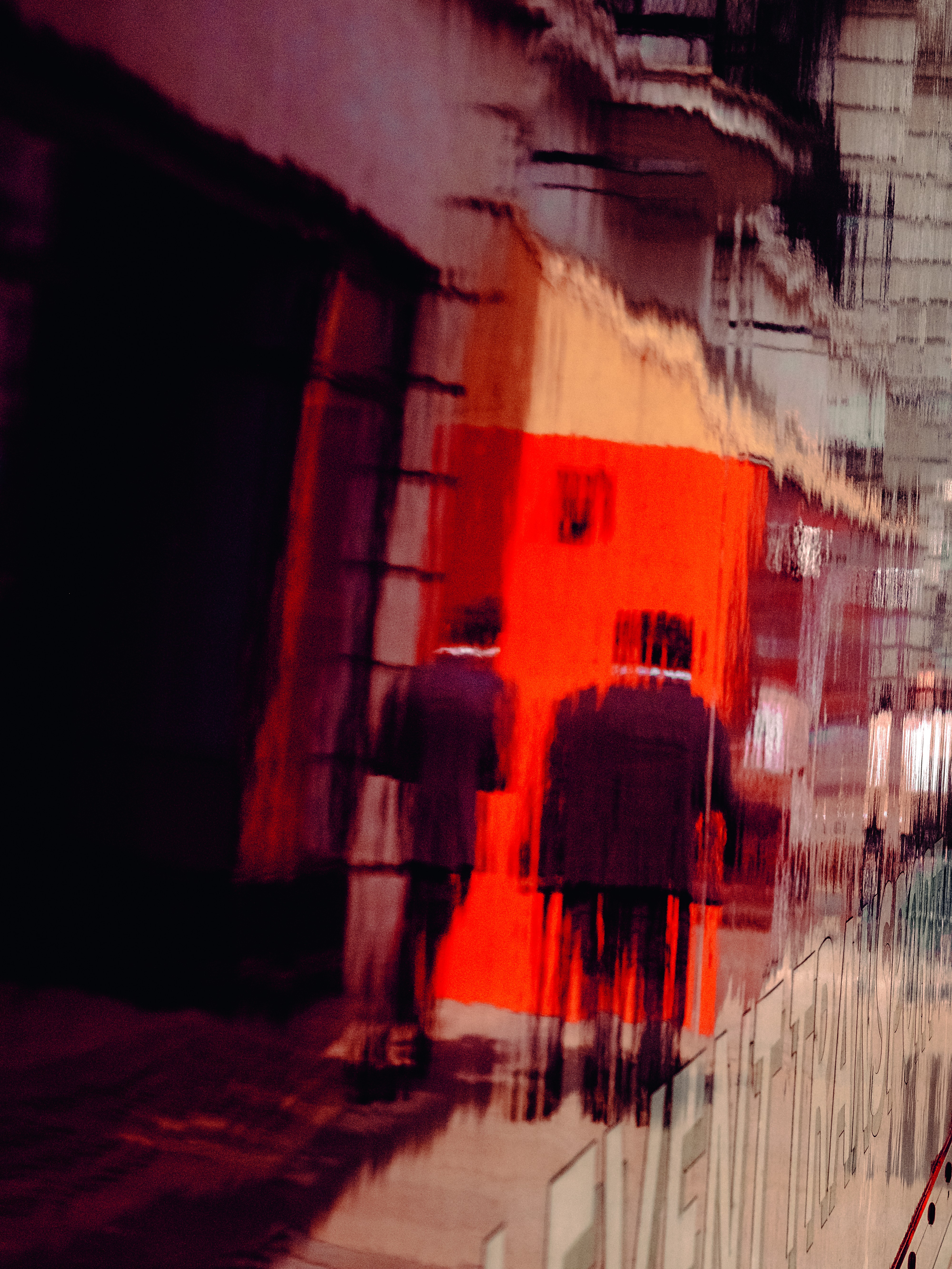 An abstract, motion-blurred street photograph in deep reds and blacks showing two ghostly figures walking past a vivid orange-red building facade.