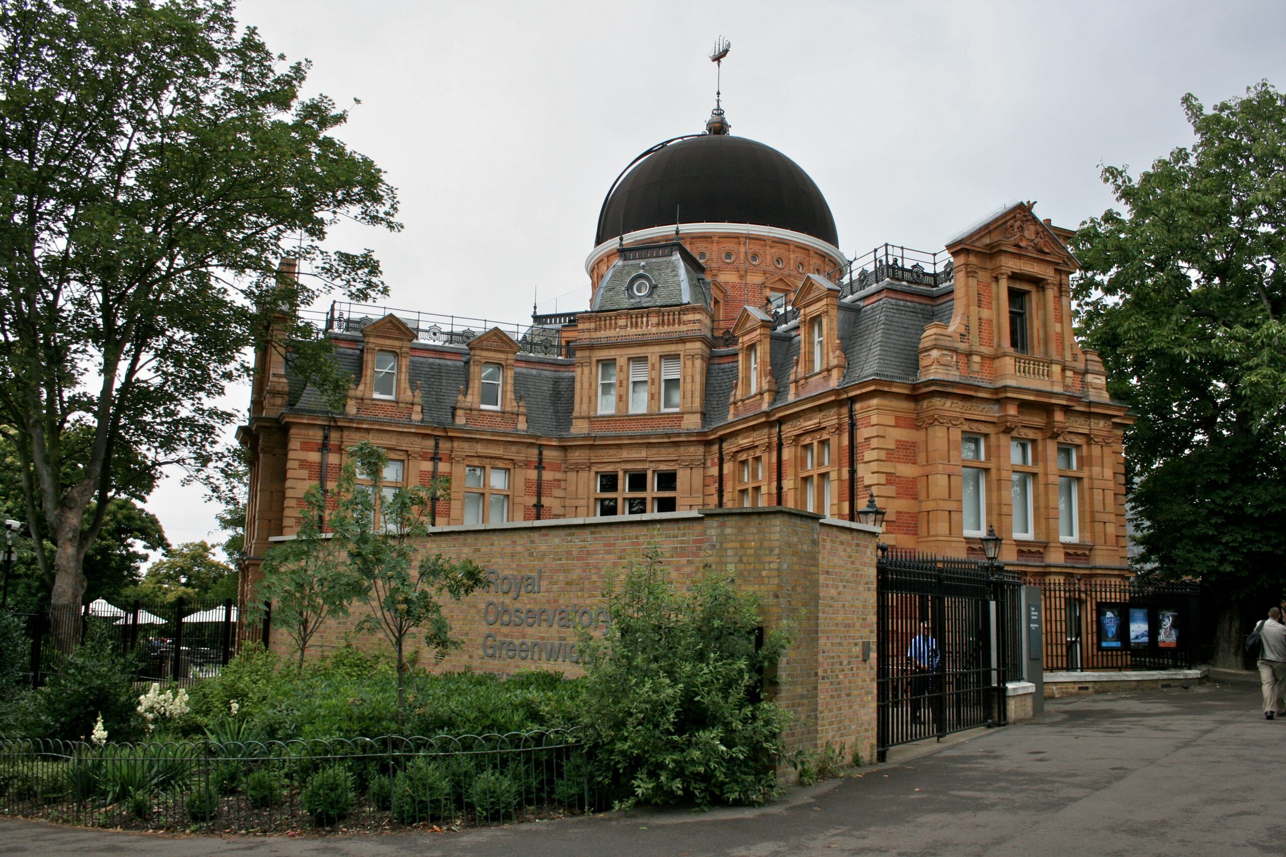A large, multi-story brick building features ornate stone trimming, pitched grey roofs, and a massive dark dome crowning its center. In the foreground, a tan brick wall partially hides the structure, surrounded by green trees, bushes, and a small garden enclosed by a black metal fence. The scene is shot from a slightly low angle on a paved path under an overcast, pale sky.
