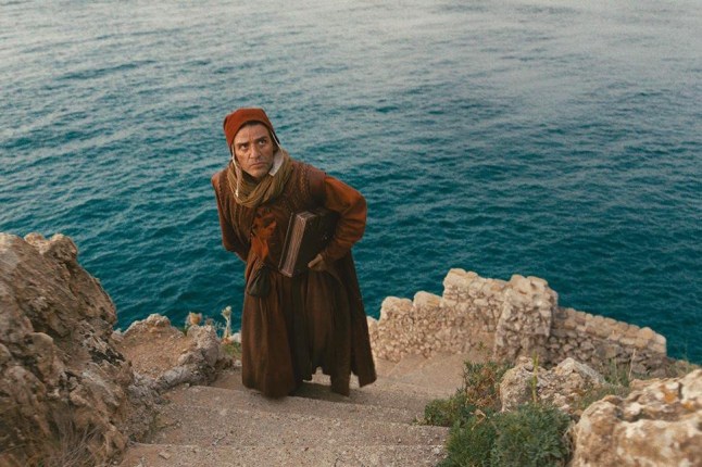 Oscar Isaac as Dante Alighieri in red robes climbing steps on mountain with books under his arm in In the Hand of Dante