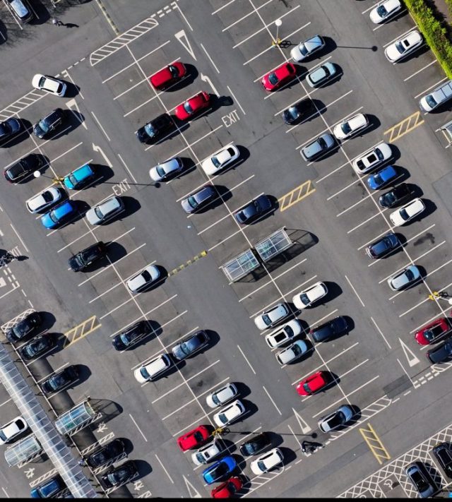 Aerial drone view of multiple coloured cars and vehicles parked in white painted spaces in a large town centre pay and display car park in UK