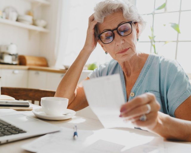 Indoor image of thoughtful puzzled senior woman with gray hair in glasses looking at paycheck holding head in hand, upset with high rent charge, sitting at kitchen table in front of laptop
