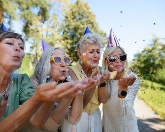 Senior female friends with party hats and sunglasses having celebration outdoors in garden.