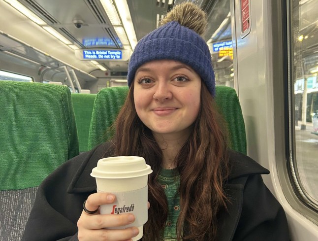 Lauren Jeffries on a train, holding a takeaway hot drink and smiling (Picture: Lauren Jeffries)