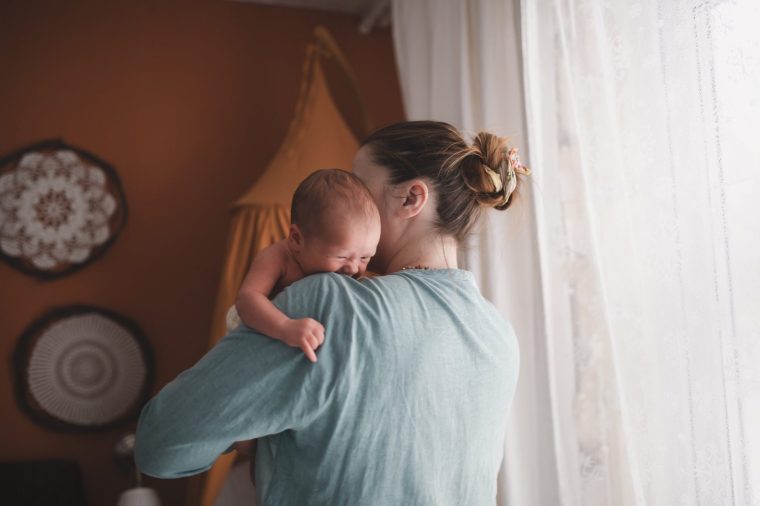 Mother with crying newborn baby at cradle of house. Terracotta and mustard color. Natural reusable diapers. Side view of new mother comforting her newborn while crying.