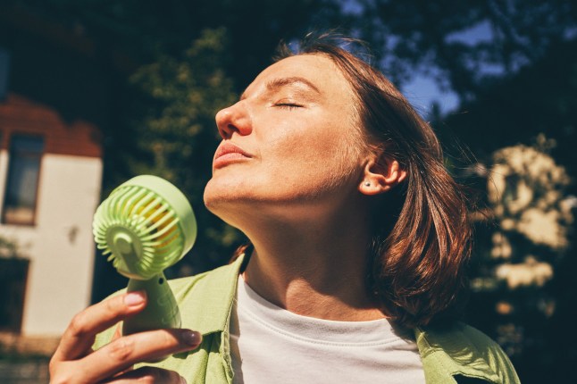 A relaxed woman holds a green portable fan outdoors on a sunny day, enjoying the breeze.