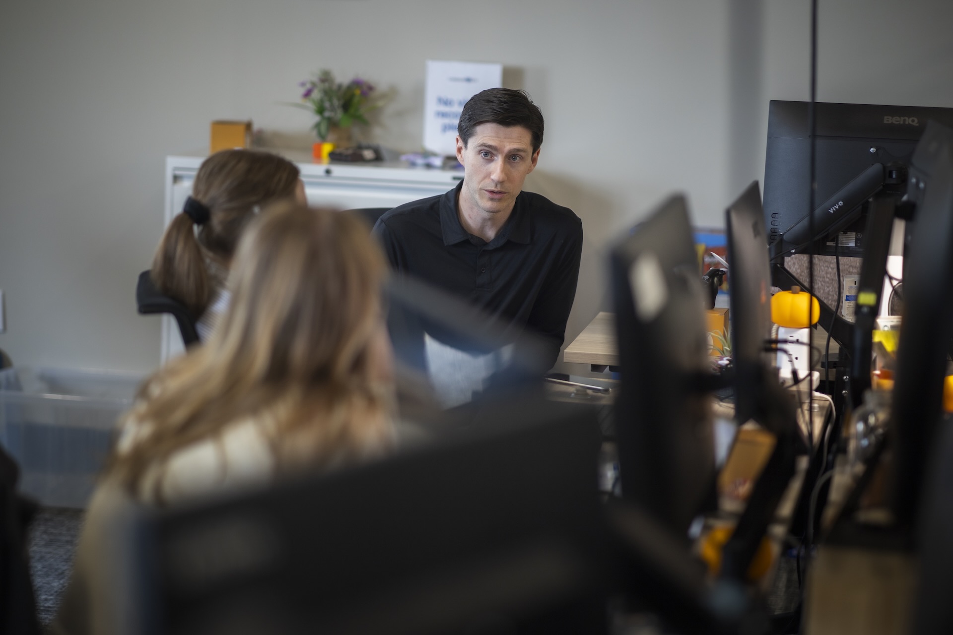 A man wearing a dark shirt sits in front of two people, foregrounded, in an office. He appears to be speaking with them.