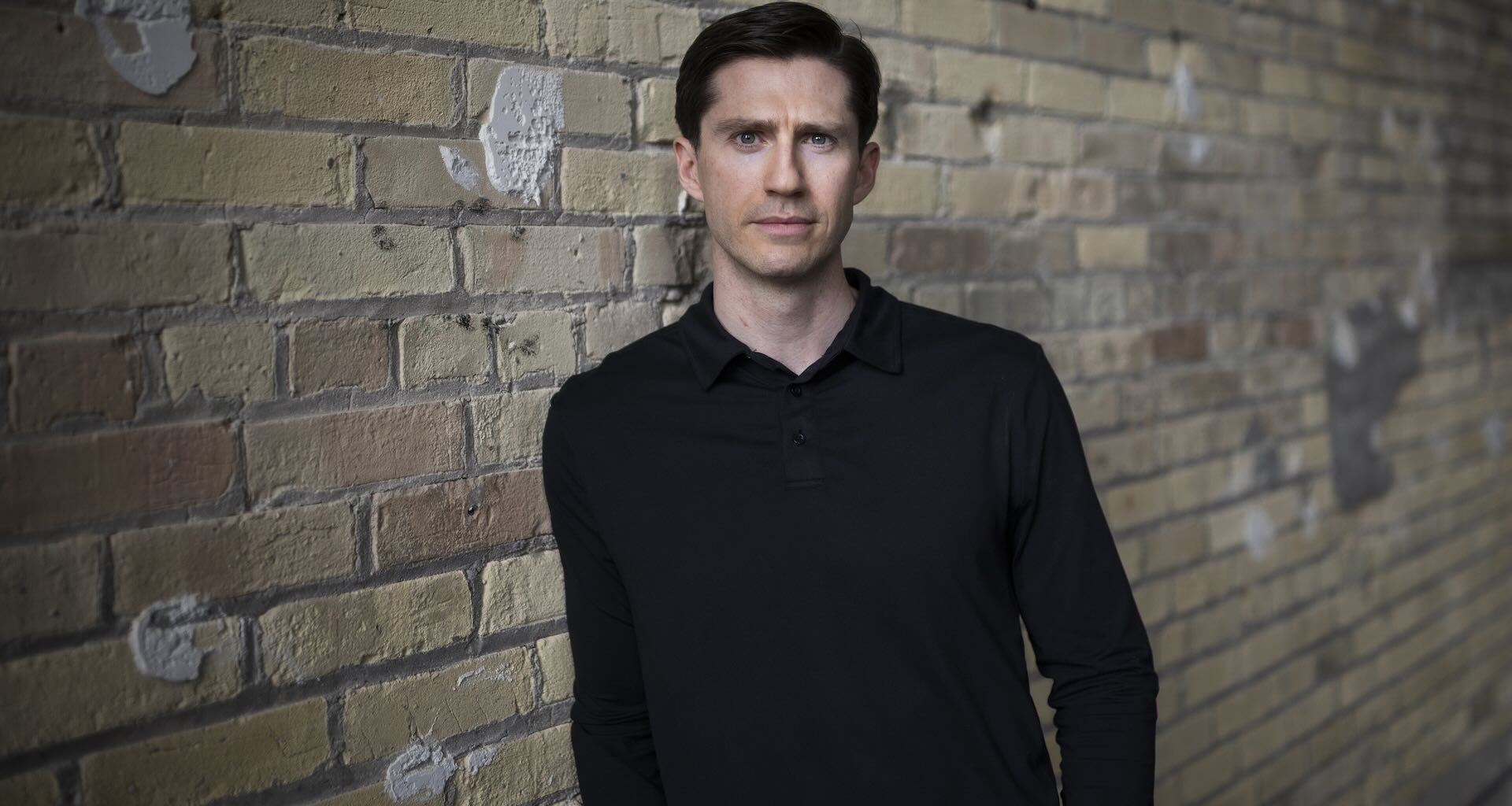 A man wearing a dark shirt is pictured against a brick wall. He is looking directly into the camera. with a serious facial expression.