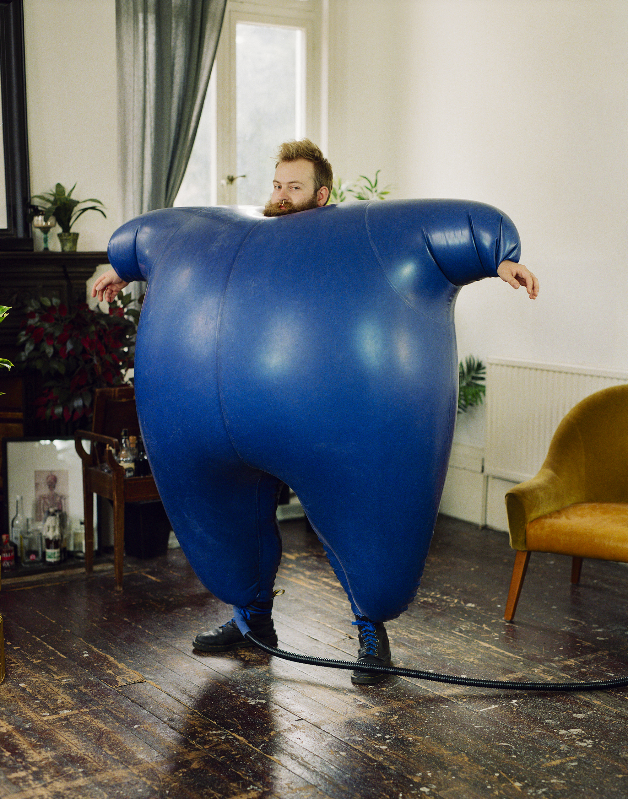 A bearded man peers over the top of an inflated blue latex body suit that engulfs his torso, standing on a wooden floor in a domestic interior.