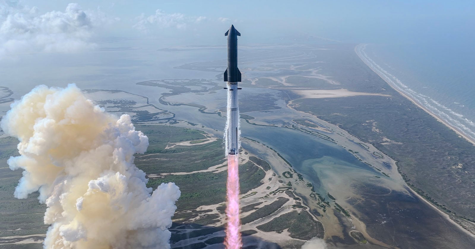 A rocket launches into the sky above a coastal landscape, leaving a trail of smoke and flames behind as it ascends over water and marshland.