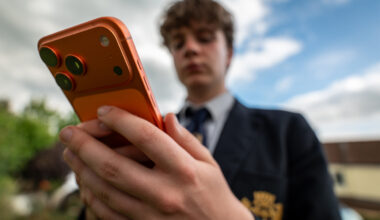 A teenager in a school uniform holding an orange iPhone out in front of them.