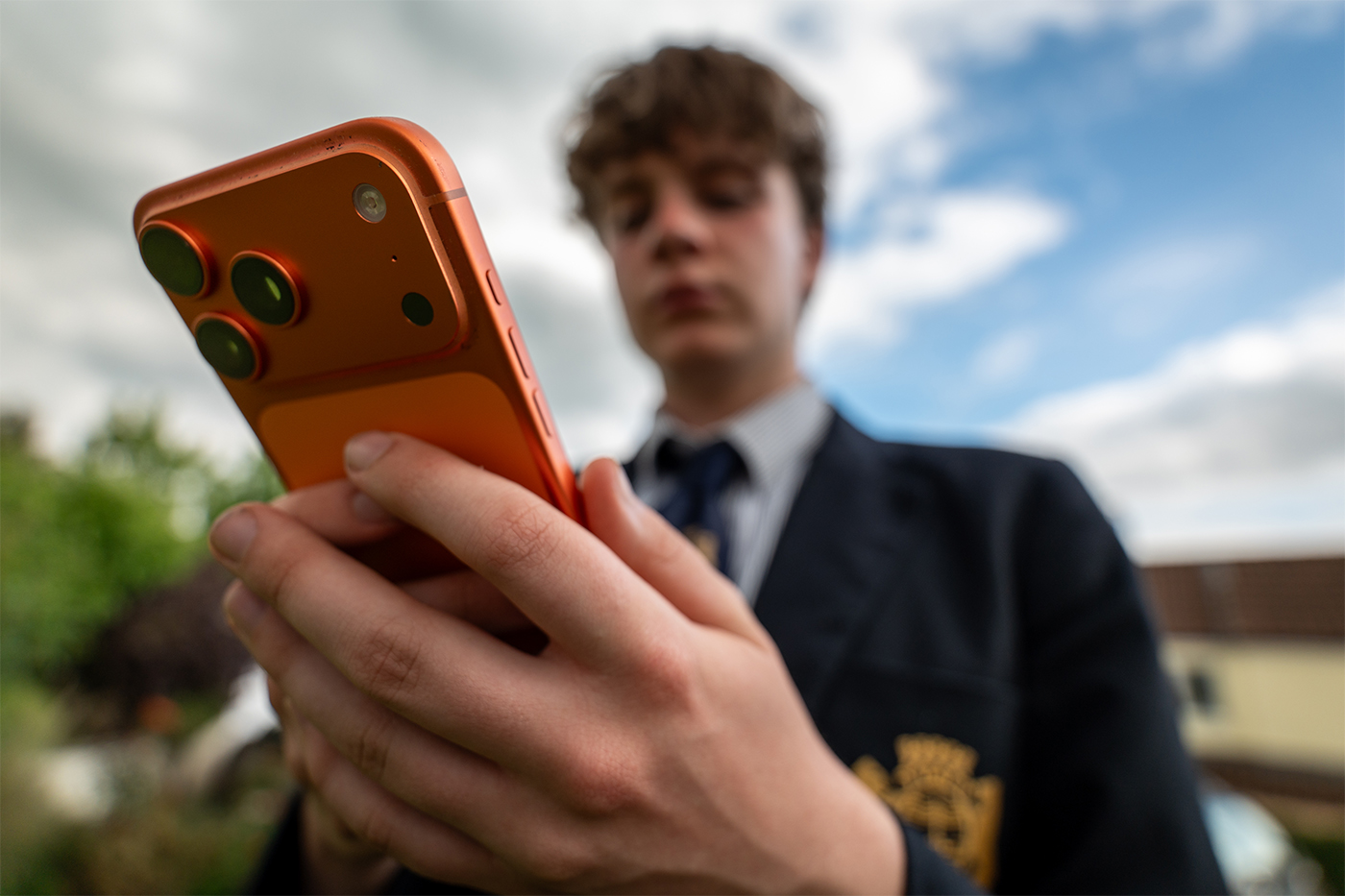 A teenager in a school uniform holding an orange iPhone out in front of them.