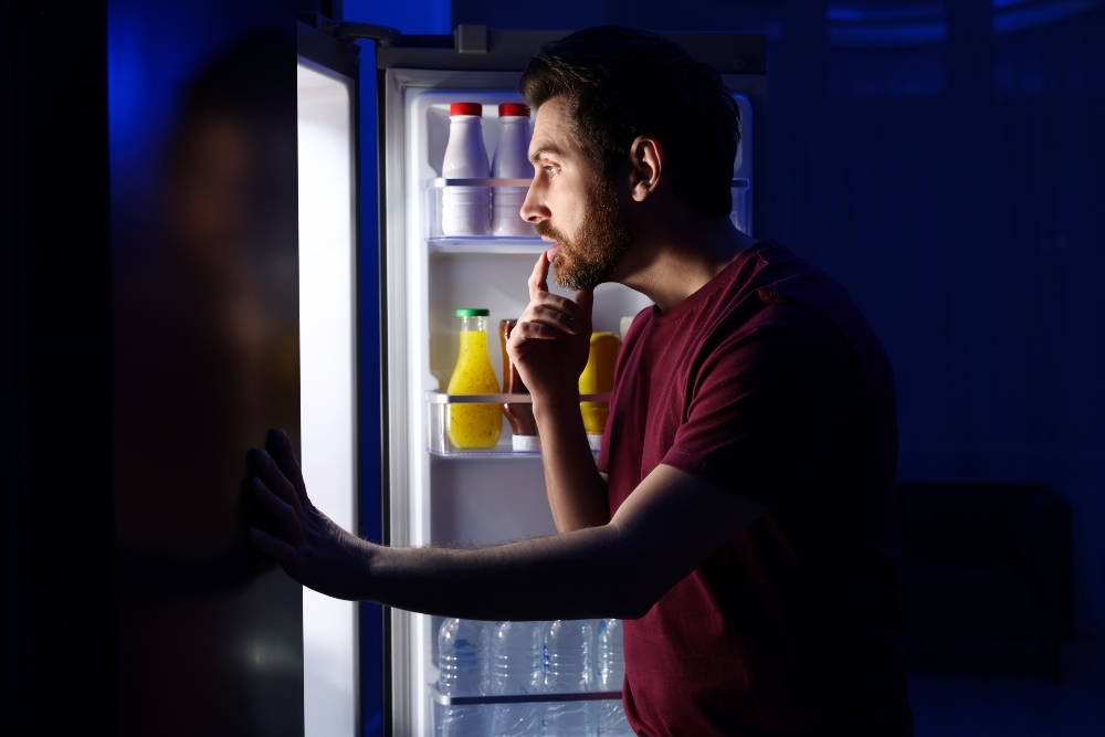 Shot of man opening the fridge in the dark looking for a late-night snack, to illustrate article about meal timing health benefits