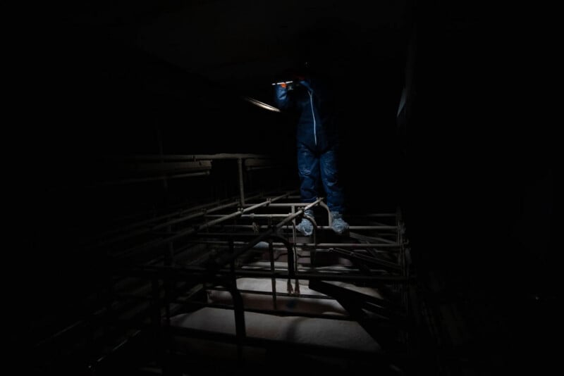 A person in a blue protective suit stands on metal beams in a dark, industrial setting, illuminated by a flashlight.