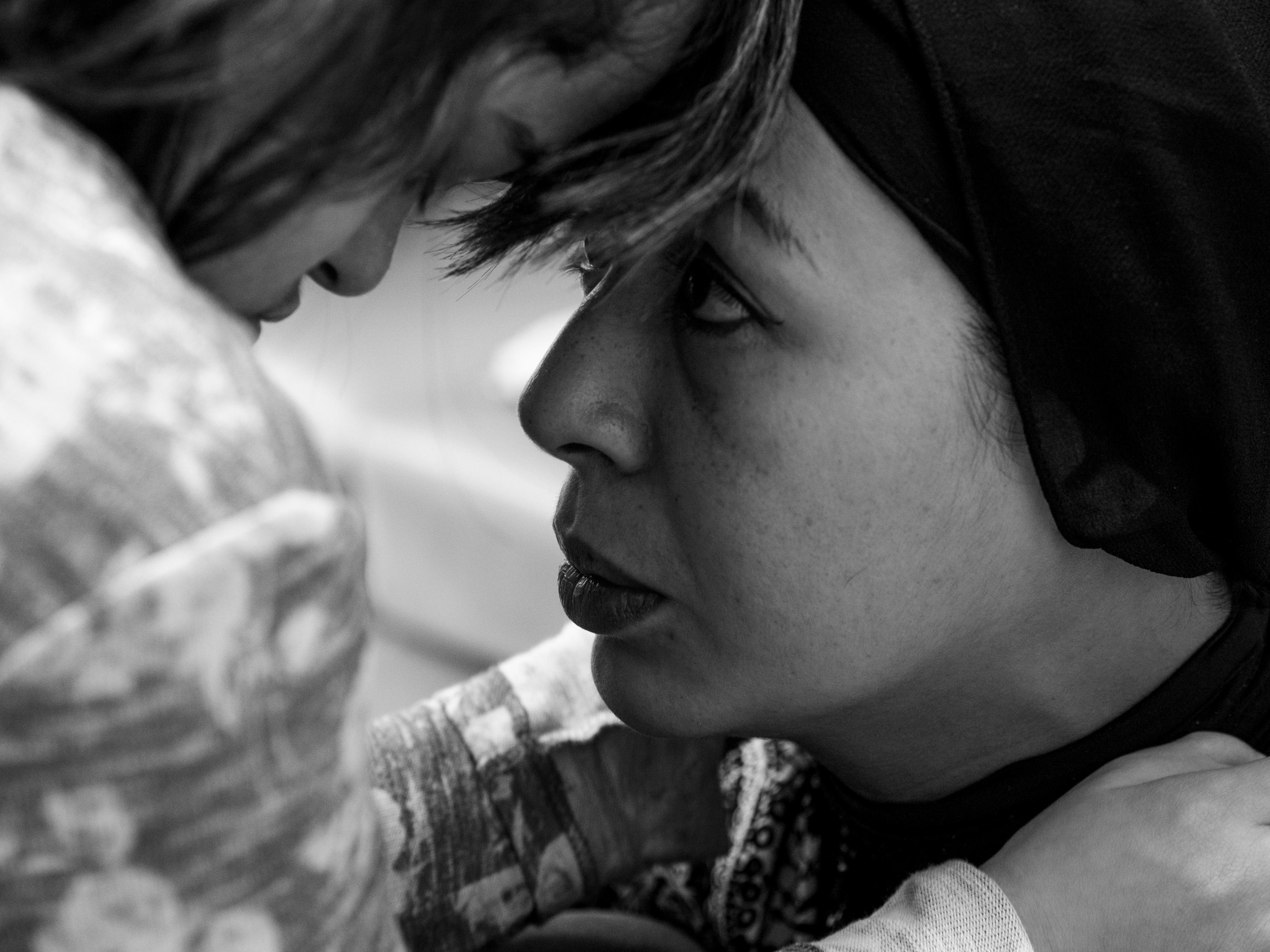 Annalisa bows her head close to Maria's in a tender moment of connection, both faces in sharp profile against a blurred outdoor background.