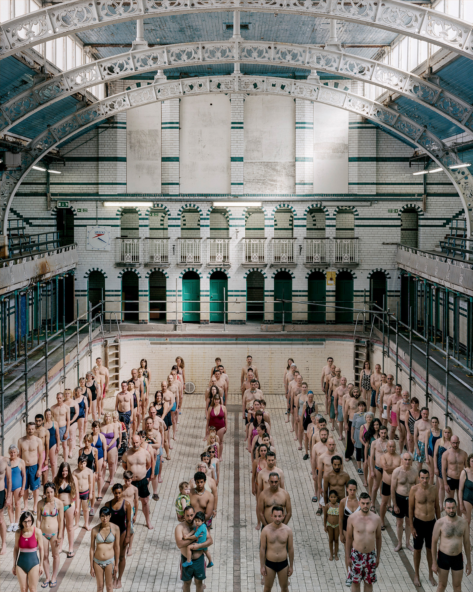 Over a hundred swimmers in swimwear stand in rows across the empty tiled floor of a grand Victorian public baths, looking towards the camera.