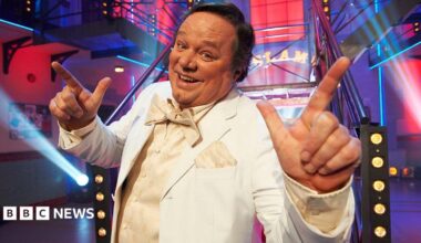 Ted Robbins in a cream tuxedo, waistcoat ad bow tie in a brightly lit set with spotlights. He has short dark hair and is smiling at the camera with his hands raised and index fingers pointing in the air