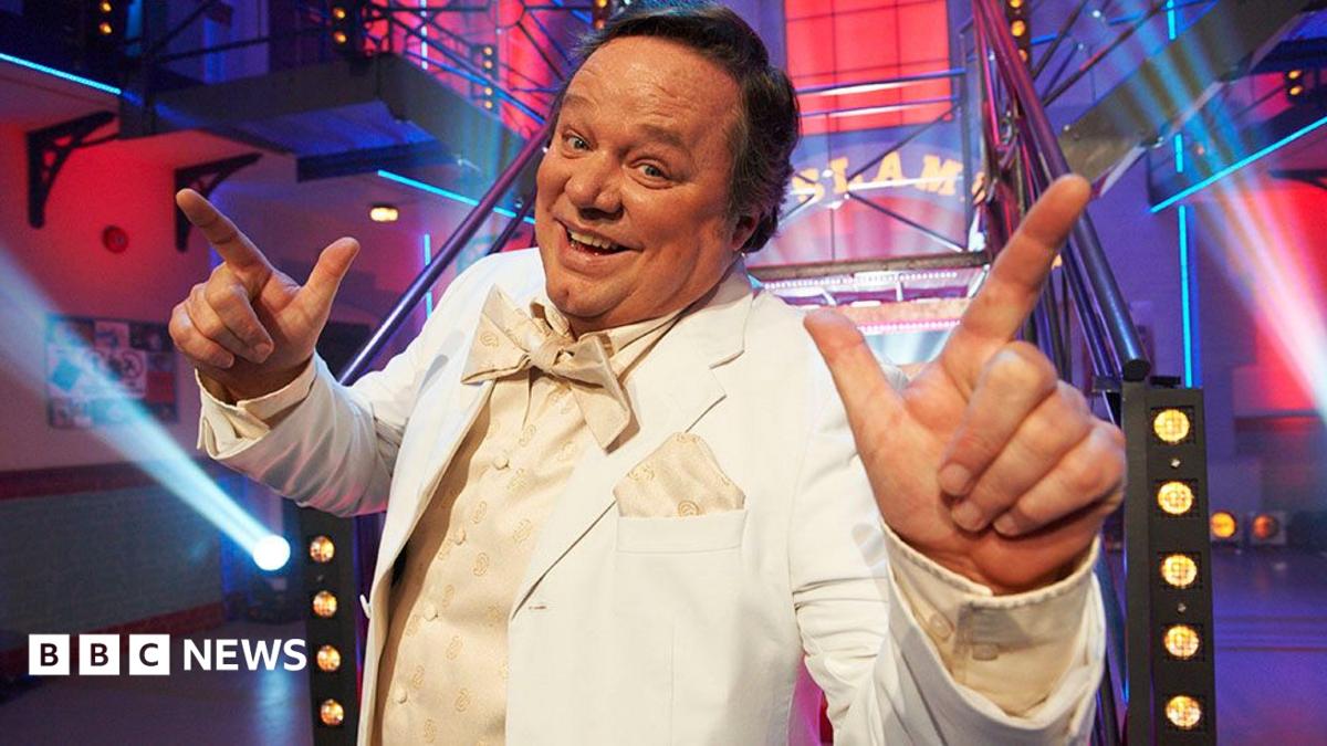 Ted Robbins in a cream tuxedo, waistcoat ad bow tie in a brightly lit set with spotlights. He has short dark hair and is smiling at the camera with his hands raised and index fingers pointing in the air