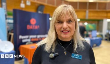 Helen Wylde-Chamberlain, CEO of Devon Chamber of Commerce, wearing a black sweater with a silver necklace. She has a small microphone on the left and a name tag on the right.