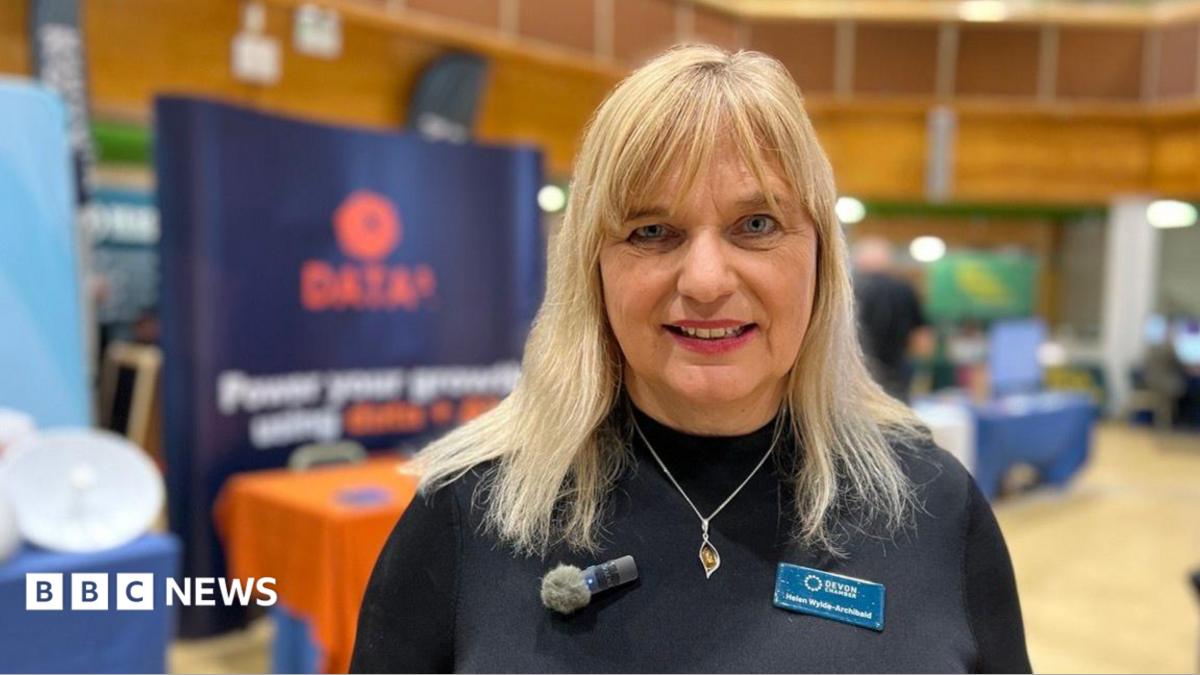 Helen Wylde-Chamberlain, CEO of Devon Chamber of Commerce, wearing a black sweater with a silver necklace. She has a small microphone on the left and a name tag on the right.